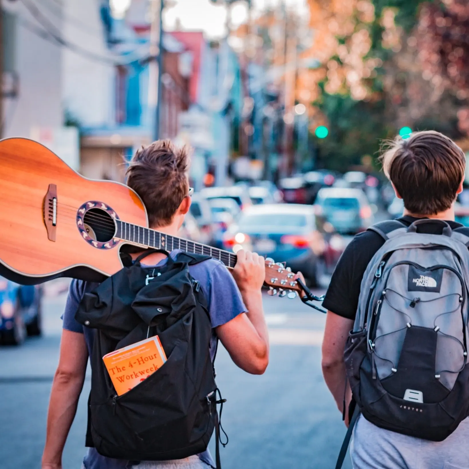 two men walking down the street with backpacks on. One has a guitar over his shoulder.