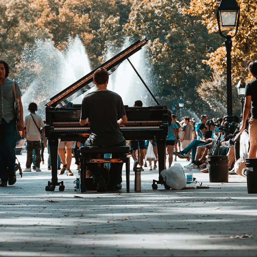 Male piano player sitting in the center of washington square park with trees overhanging and the fountain in front.