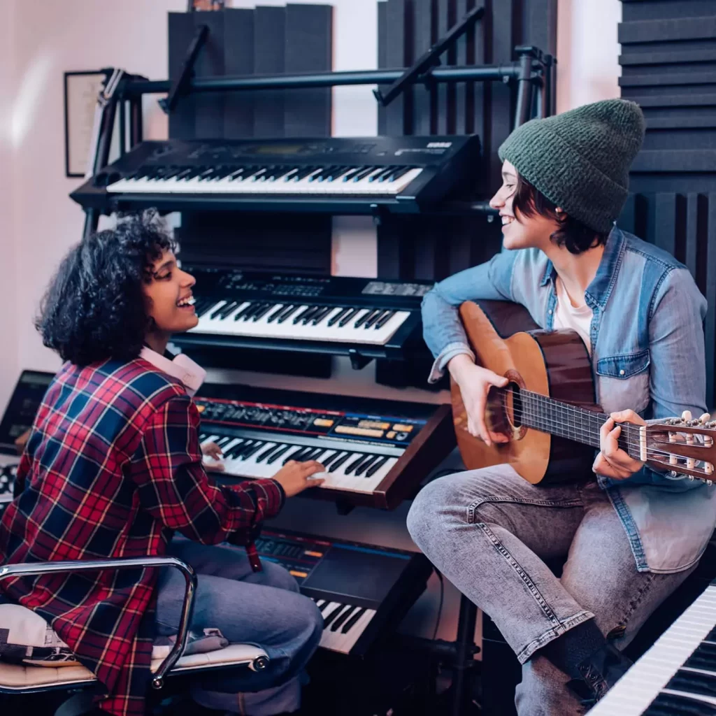 Two musicians in a studio, one playing keyboard and the other guitar, smiling and collaborating on music.