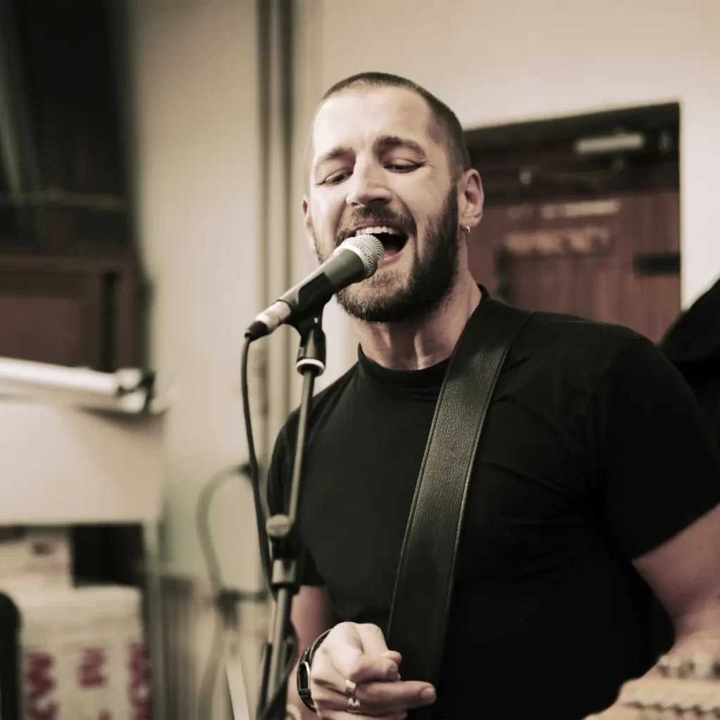 Bearded musician passionately singing into a microphone while playing guitar indoors.