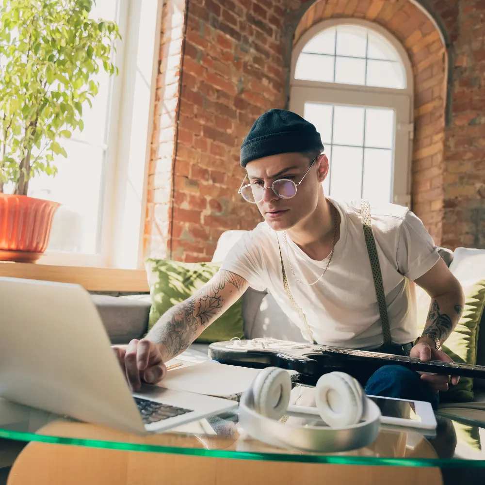 A musician with a guitar and laptop, preparing a Spotify playlist submission in a bright loft-style room.